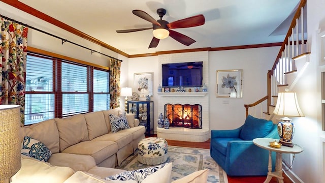 living room featuring a ceiling fan, stairway, wood finished floors, crown molding, and a brick fireplace