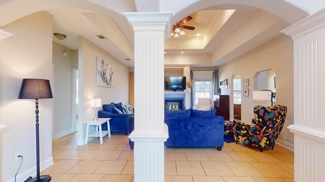 tiled living room with ceiling fan, ornamental molding, a raised ceiling, and decorative columns