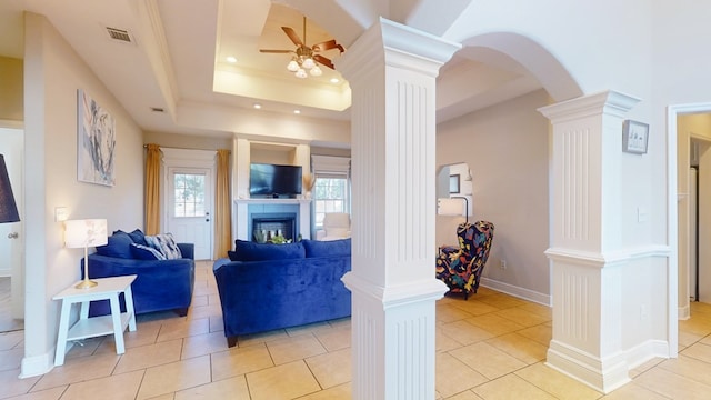 living room featuring crown molding, light tile patterned floors, a raised ceiling, ceiling fan, and decorative columns