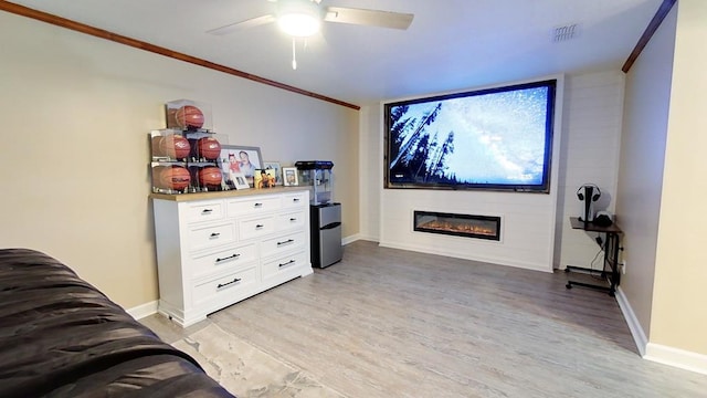 cinema with light wood-type flooring, ceiling fan, baseboards, crown molding, and a glass covered fireplace
