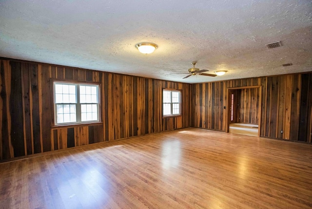 unfurnished room with a textured ceiling, visible vents, wooden walls, a ceiling fan, and light wood-type flooring