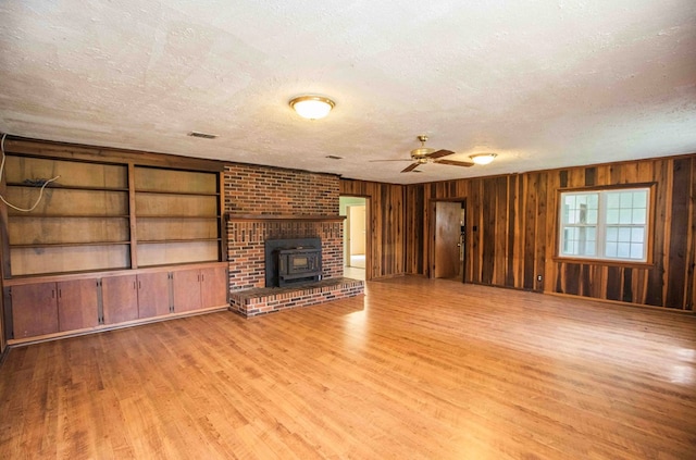 unfurnished living room with a textured ceiling, light wood-style flooring, wood walls, a ceiling fan, and a wood stove