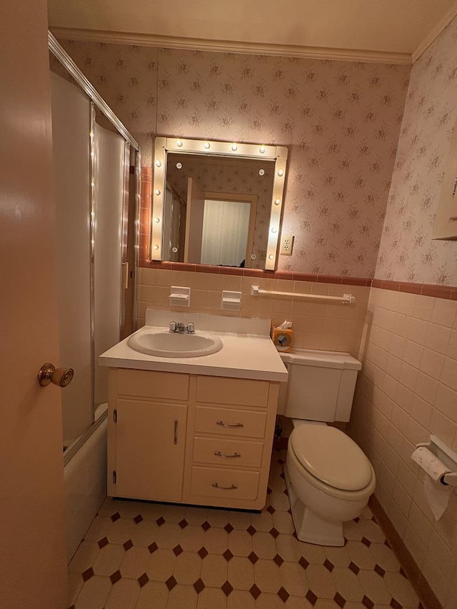 bathroom featuring wallpapered walls, a wainscoted wall, toilet, shower / bath combination with glass door, and vanity