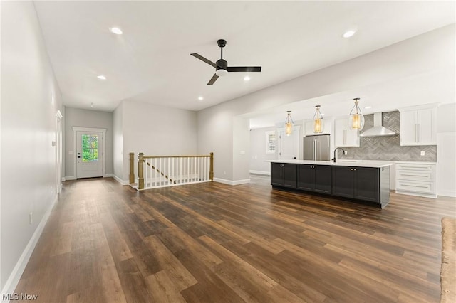 kitchen with stainless steel fridge, wall chimney exhaust hood, a kitchen island with sink, pendant lighting, and white cabinetry