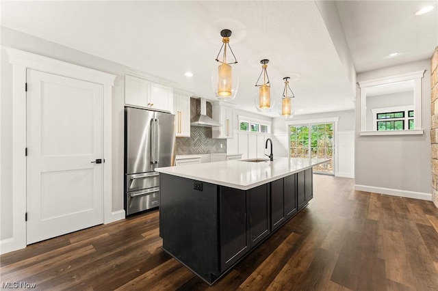 kitchen featuring high end refrigerator, wall chimney range hood, sink, hanging light fixtures, and an island with sink