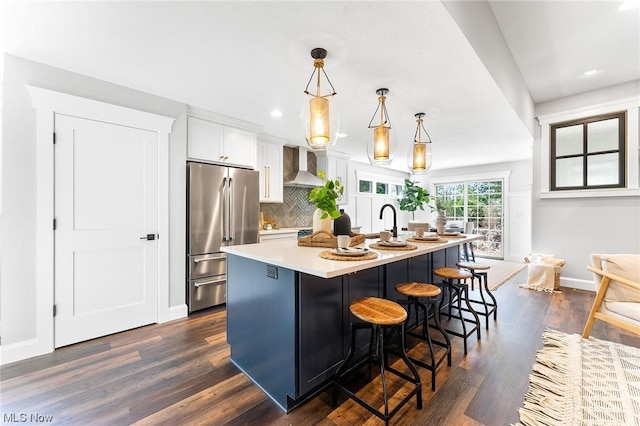 kitchen featuring wall chimney range hood, pendant lighting, a kitchen island with sink, high end fridge, and white cabinets