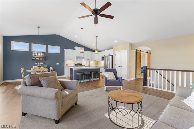 living room featuring vaulted ceiling, ceiling fan with notable chandelier, sink, light hardwood / wood-style floors, and a barn door