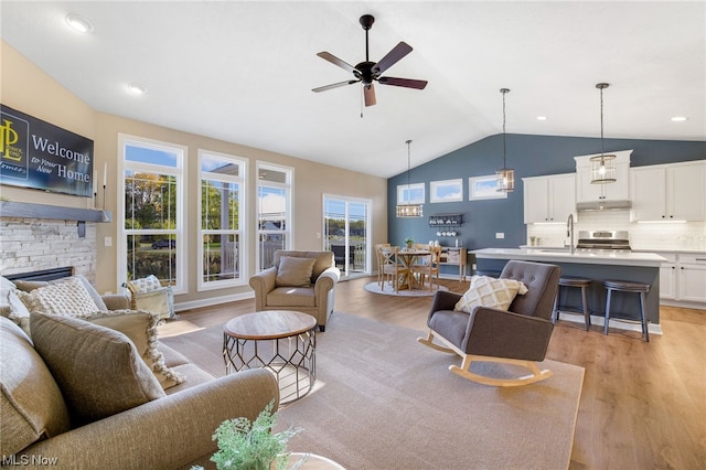 living room with sink, vaulted ceiling, light hardwood / wood-style flooring, a stone fireplace, and ceiling fan