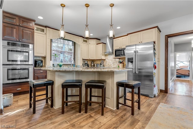 kitchen with appliances with stainless steel finishes, a center island with sink, decorative backsplash, light wood-type flooring, and wall chimney exhaust hood