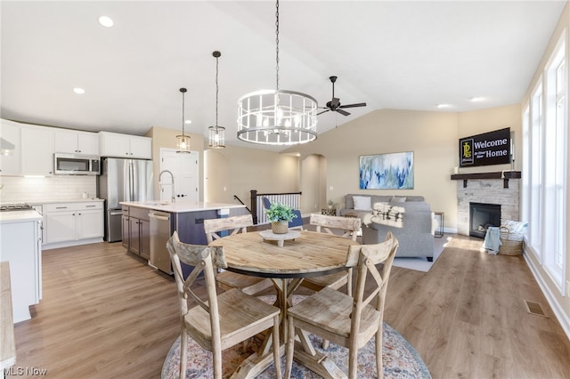 dining space featuring ceiling fan with notable chandelier, a stone fireplace, sink, light hardwood / wood-style flooring, and vaulted ceiling