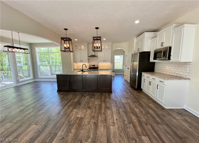 kitchen featuring wall chimney exhaust hood, white cabinetry, appliances with stainless steel finishes, and an island with sink