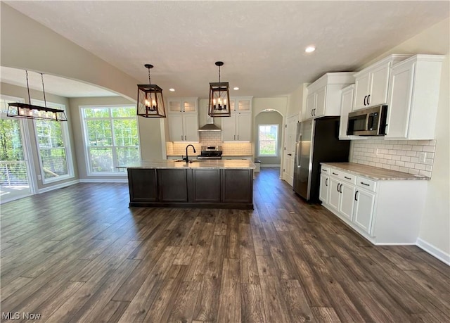 kitchen featuring a kitchen island with sink, stainless steel appliances, white cabinets, hanging light fixtures, and wall chimney range hood