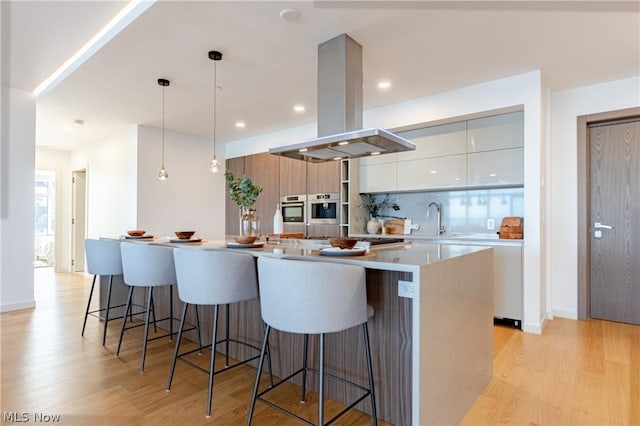 kitchen featuring white cabinetry, a center island with sink, light wood-type flooring, pendant lighting, and island exhaust hood