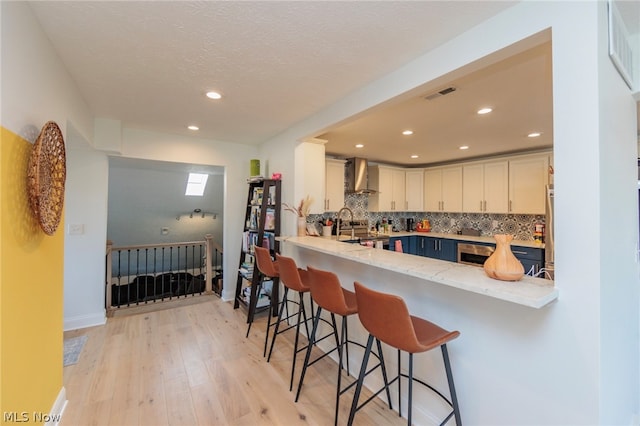 kitchen with white cabinetry, wall chimney range hood, kitchen peninsula, a breakfast bar area, and light hardwood / wood-style floors