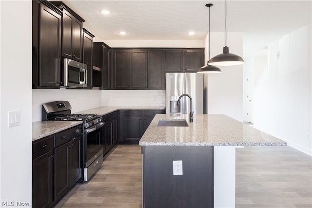 kitchen with appliances with stainless steel finishes, hanging light fixtures, light hardwood / wood-style floors, light stone countertops, and a center island with sink