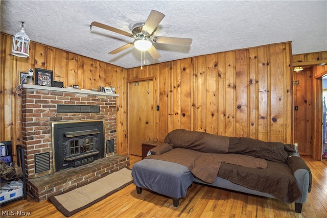 living room featuring a brick fireplace, ceiling fan, light hardwood / wood-style floors, a textured ceiling, and wooden walls