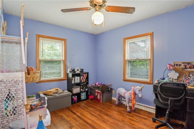 recreation room featuring a healthy amount of sunlight, wood-type flooring, ceiling fan, and a baseboard heating unit