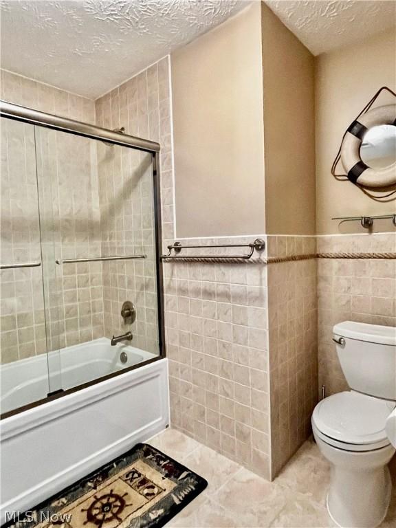 bathroom featuring tile walls, a textured ceiling, bath / shower combo with glass door, and toilet