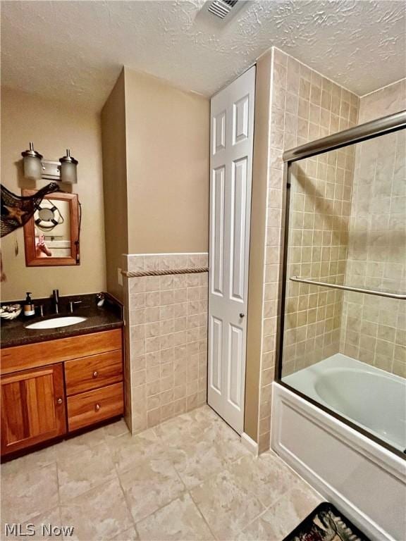 bathroom with bath / shower combo with glass door, tile walls, vanity, and a textured ceiling