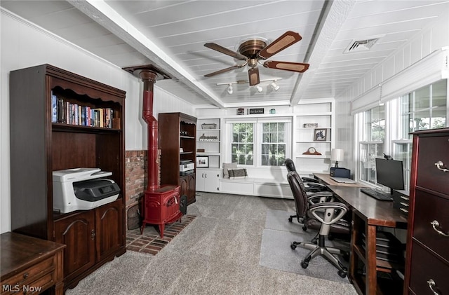 carpeted home office featuring a wealth of natural light, visible vents, beamed ceiling, and a wood stove