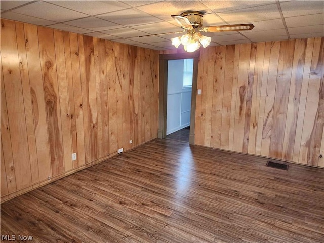 empty room with a drop ceiling, dark wood-type flooring, wooden walls, and ceiling fan