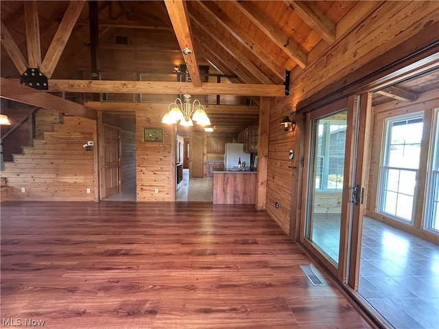 unfurnished living room with an inviting chandelier, wood-type flooring, wooden ceiling, wooden walls, and beamed ceiling