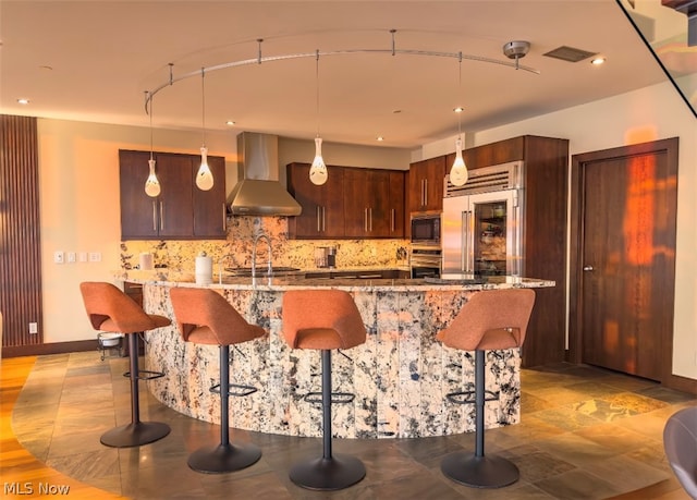 kitchen featuring backsplash, decorative light fixtures, wall chimney exhaust hood, and built in appliances