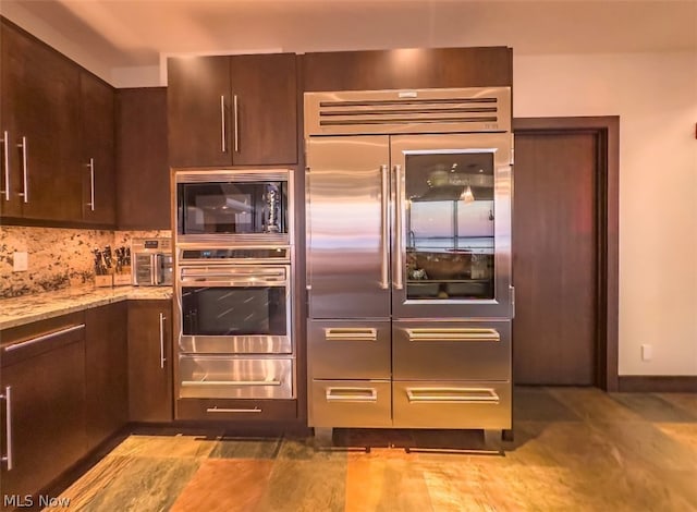 kitchen featuring built in appliances, light stone counters, dark brown cabinetry, decorative backsplash, and tile patterned floors