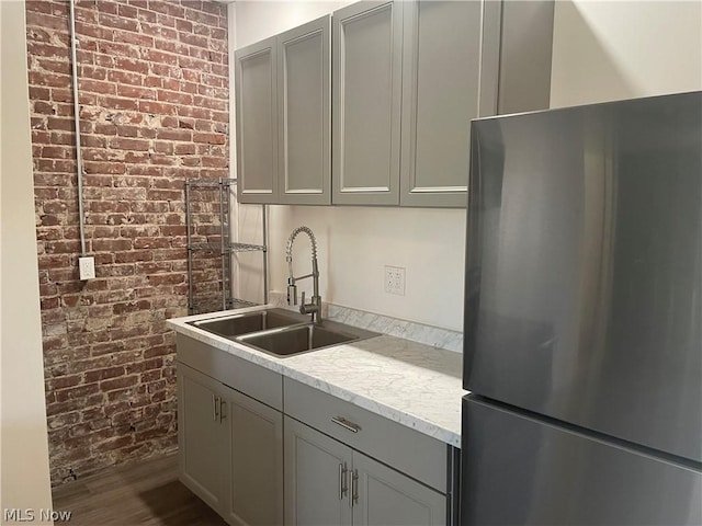 kitchen with sink, gray cabinets, stainless steel refrigerator, light stone countertops, and brick wall