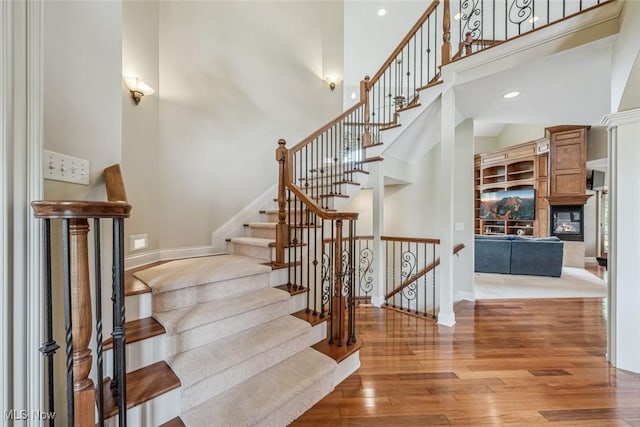 staircase with a glass covered fireplace, recessed lighting, wood-type flooring, baseboards, and a towering ceiling