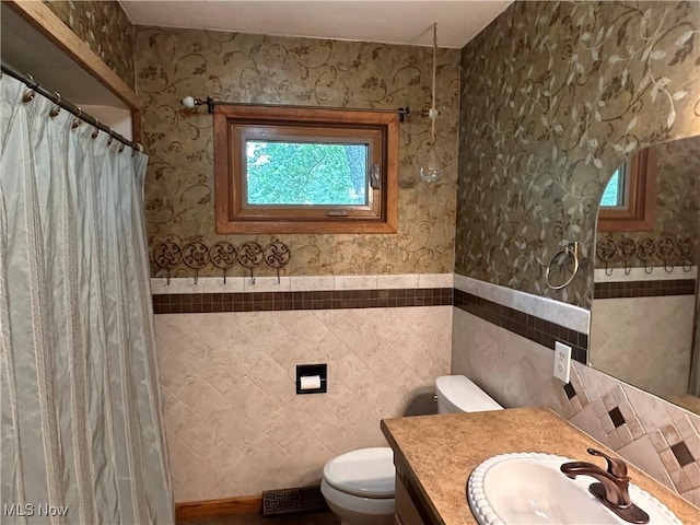 bathroom with wallpapered walls, tile walls, a wainscoted wall, toilet, and vanity