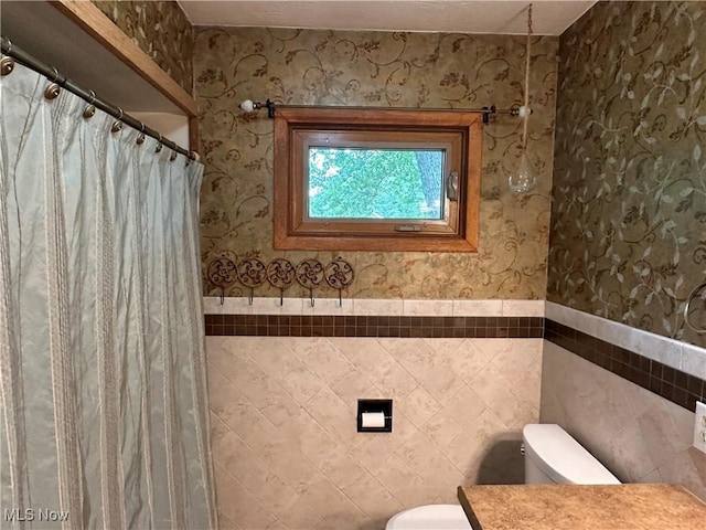 bathroom with wainscoting, toilet, wallpapered walls, and tile walls