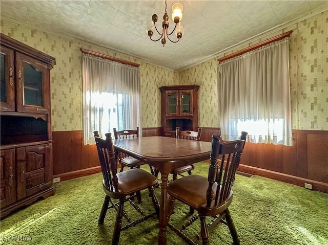 carpeted dining space with wallpapered walls, crown molding, a chandelier, and a wainscoted wall