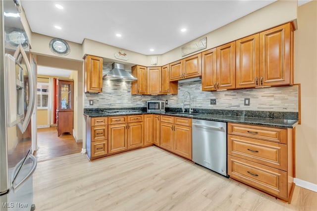 kitchen with wall chimney range hood, stainless steel appliances, dark stone countertops, light wood-type flooring, and decorative backsplash