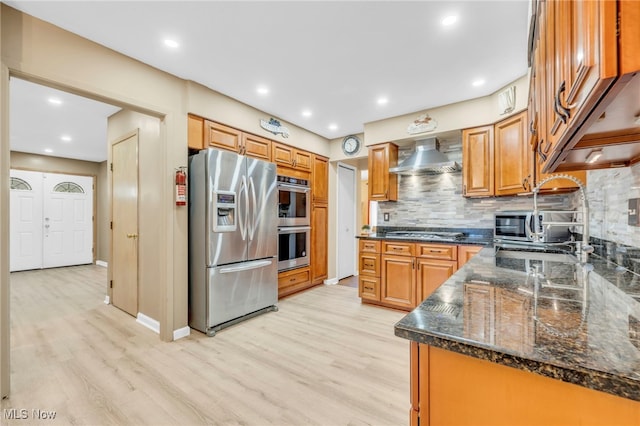 kitchen featuring appliances with stainless steel finishes, light hardwood / wood-style floors, dark stone countertops, tasteful backsplash, and wall chimney range hood