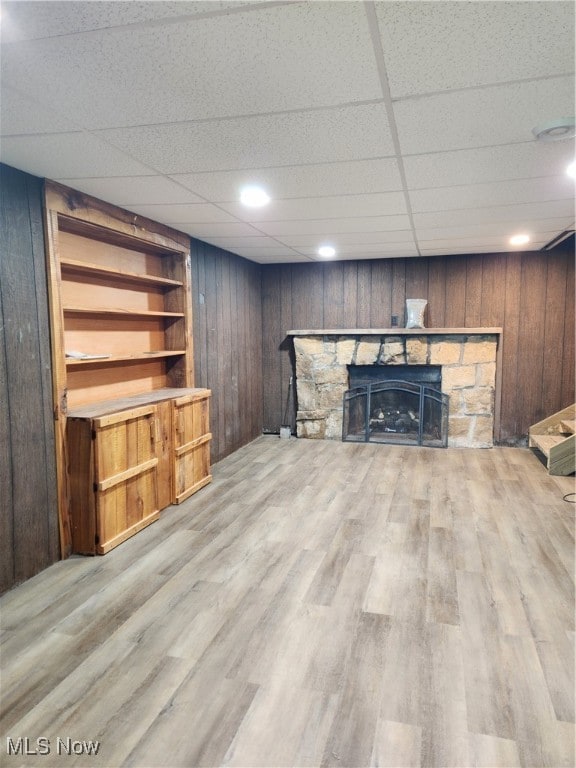 unfurnished living room with light wood-type flooring, a paneled ceiling, wooden walls, and a stone fireplace