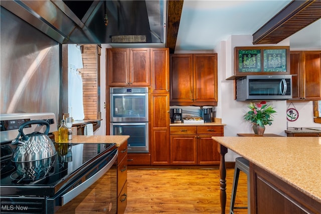 kitchen with appliances with stainless steel finishes, wall chimney range hood, light stone counters, and light hardwood / wood-style floors