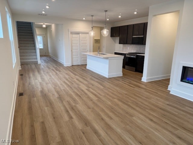 kitchen with stainless steel stove, a kitchen island with sink, dark brown cabinets, light hardwood / wood-style floors, and decorative light fixtures