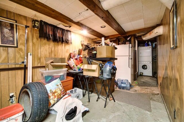 basement featuring wood walls, stacked washing maching and dryer, and white fridge