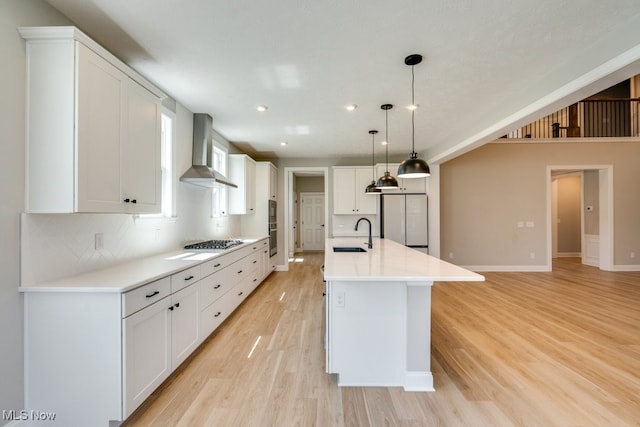 kitchen with white refrigerator, decorative light fixtures, wall chimney exhaust hood, light hardwood / wood-style floors, and a center island with sink