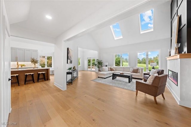 living room featuring high vaulted ceiling, light wood-style flooring, a glass covered fireplace, a skylight, and baseboards