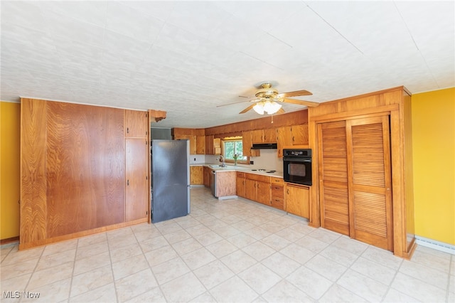 kitchen featuring wooden walls, black oven, sink, stainless steel refrigerator, and ceiling fan