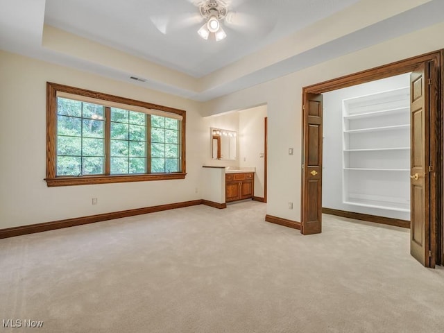 unfurnished bedroom featuring light carpet, baseboards, ensuite bathroom, a tray ceiling, and a walk in closet