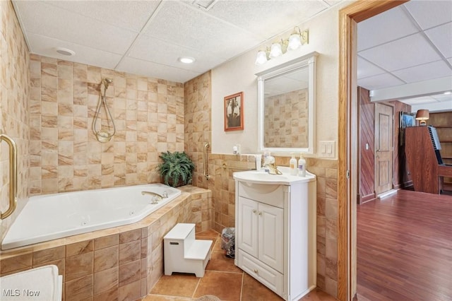 bathroom with tile walls, tile patterned flooring, vanity, a relaxing tiled tub, and a drop ceiling