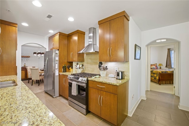 kitchen featuring an inviting chandelier, light stone counters, stainless steel appliances, wall chimney range hood, and tasteful backsplash