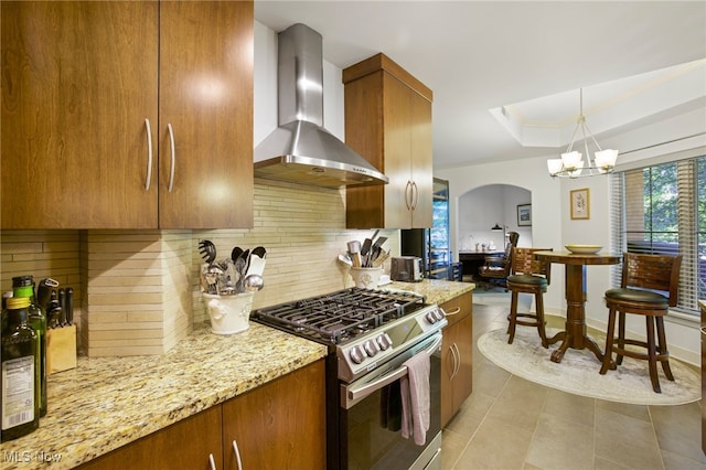 kitchen featuring stainless steel gas range, a notable chandelier, light stone counters, a tray ceiling, and wall chimney exhaust hood