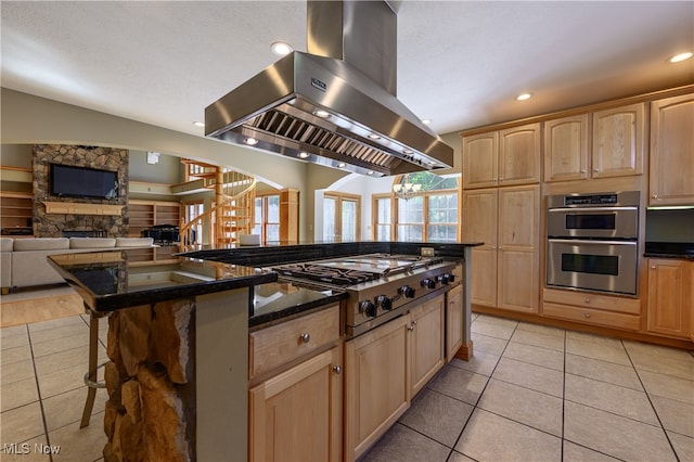 kitchen with stainless steel appliances, island range hood, light brown cabinetry, a kitchen island, and a stone fireplace