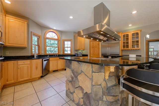 kitchen with light brown cabinetry, appliances with stainless steel finishes, island exhaust hood, a center island, and dark stone countertops