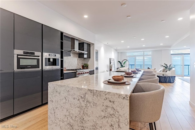 kitchen with a center island with sink, wall chimney exhaust hood, tasteful backsplash, light hardwood / wood-style floors, and stainless steel appliances