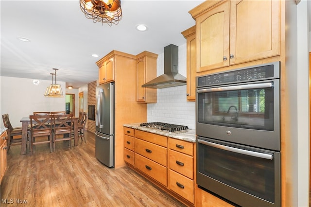 kitchen featuring decorative light fixtures, light stone countertops, light hardwood / wood-style flooring, appliances with stainless steel finishes, and wall chimney range hood